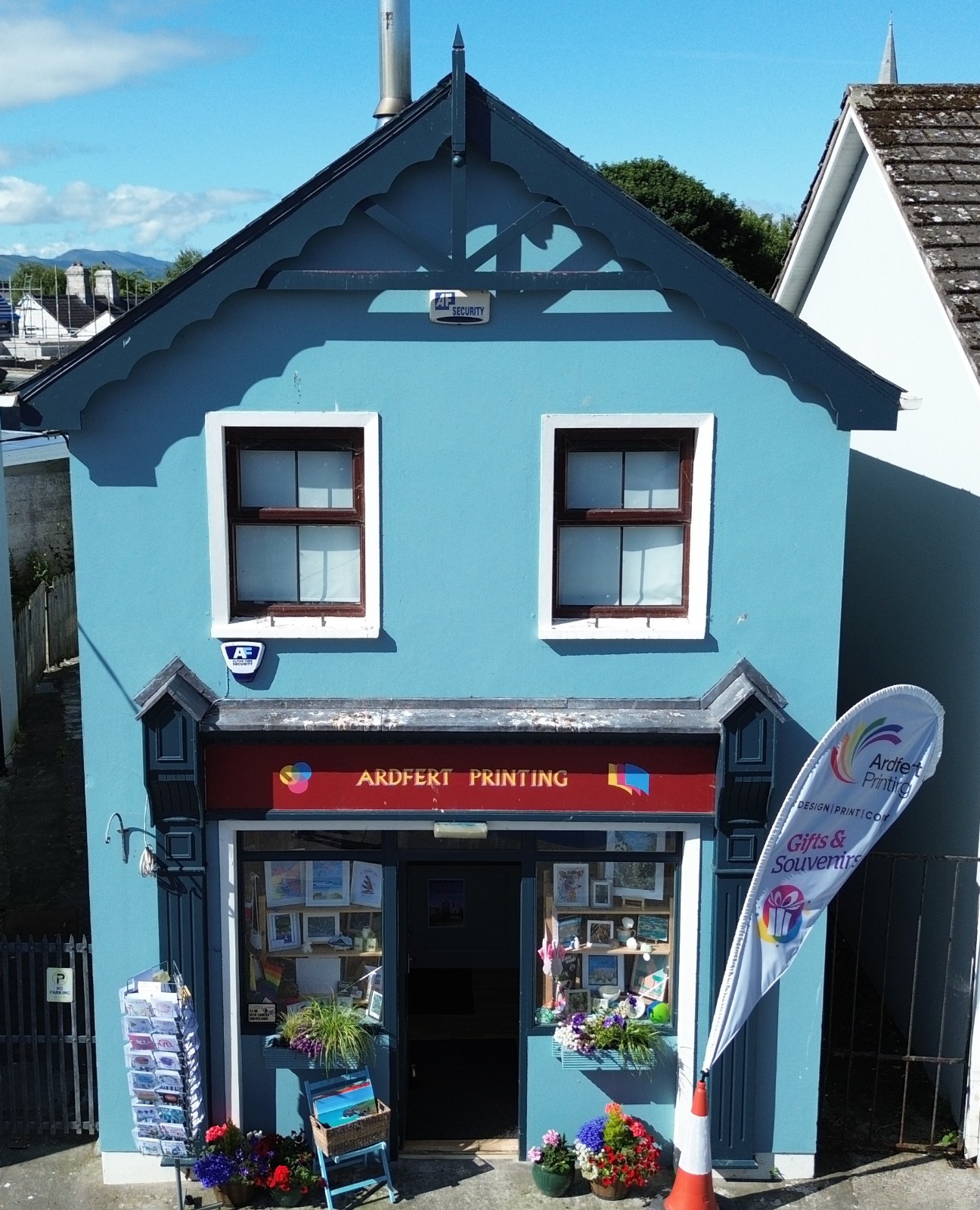 The bright blue Ardfert Printing shopfront in Ardfert Village, Co. Kerry, with window displays of Kerry prints and a Gifts & Souvenirs flag